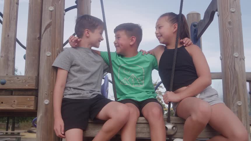 Three children two boys and a girl sit side by side on a wooden play ramp at midday holding each other and smiling warmly at one another
