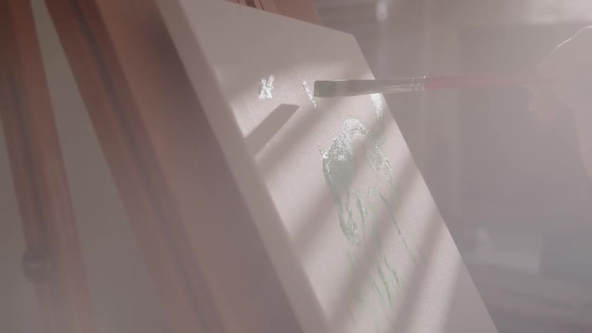 An artists arm holds a red brush painting on a canvas inside a studio at midday with sunlight streaming through the window into the room