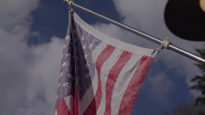 A closeup shot captures the lower portion of the flag of the united states of america hanging from a silver metal pole visible against a partly cloudy blue sky with the stars and stripes subtly lit