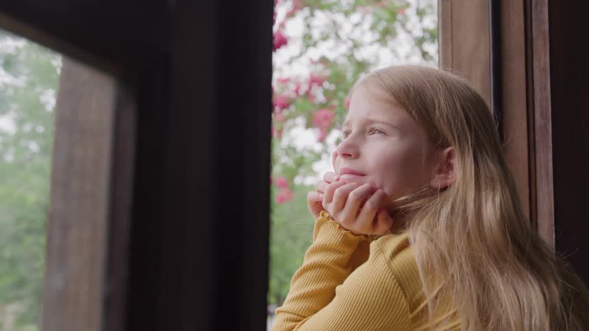The face of a little blonde girl wearing a yellow tshirt looking out of a window