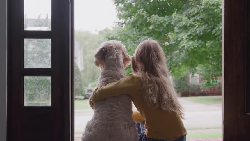 Girl sits on her doorstep with her arms around her dog and watches a lawnmower in a neighbors yard