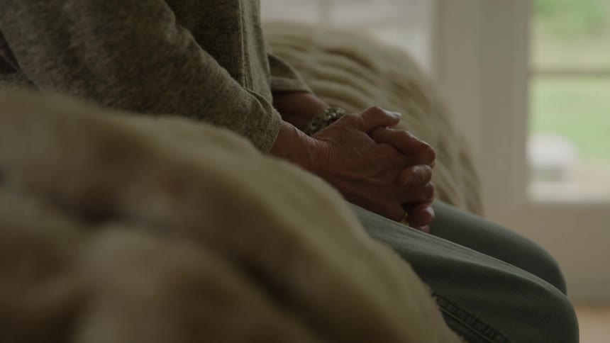 Hands clasped together resting on lap in residential setting with natural light