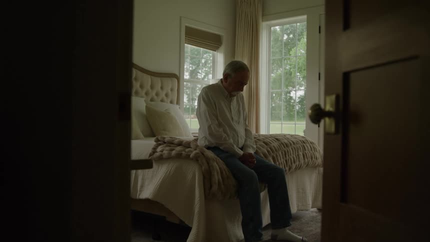 An elderly man praying while sitting at the edge of a bed in a bedroom during daytime