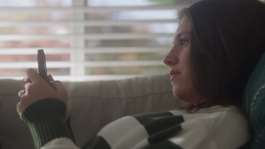 Person in white and green striped sweater reclining on couch using smartphone during daytime
