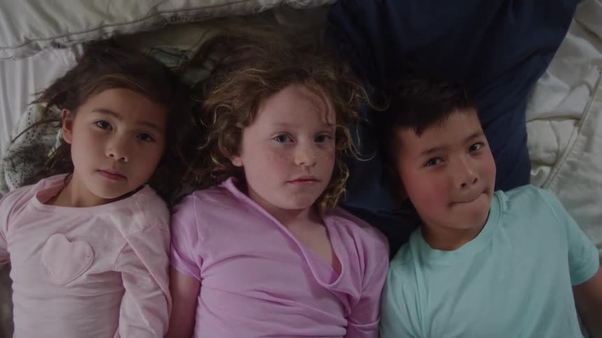 Two little girls and a little boy are lying on a bed looking ahead then they start looking at each other and laughing