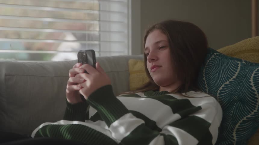 A young woman lies on a couch at midday wearing a sweatshirt scrolling through her mobile phone