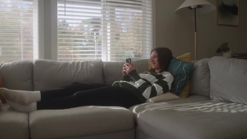 Woman lying on gray sofa using phone in naturally lit living room