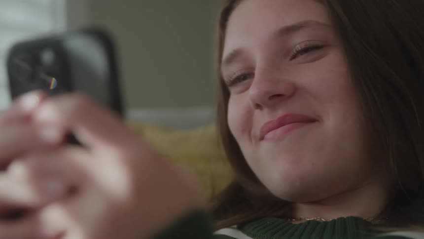 A teenage girl lies on a couch indoors using her mobile device smiling as she interacts with it