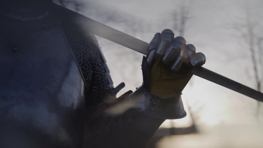 A dramatic closeup focuses on a metallic gauntlet gripping the steel of a sword with the statue and weapon heavily silhouetted and backlit by a bright hazy outdoor light