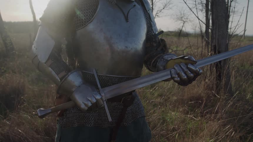 A medieval knight in full plate armor and chainmail stands ready in a field holding a large broadsword