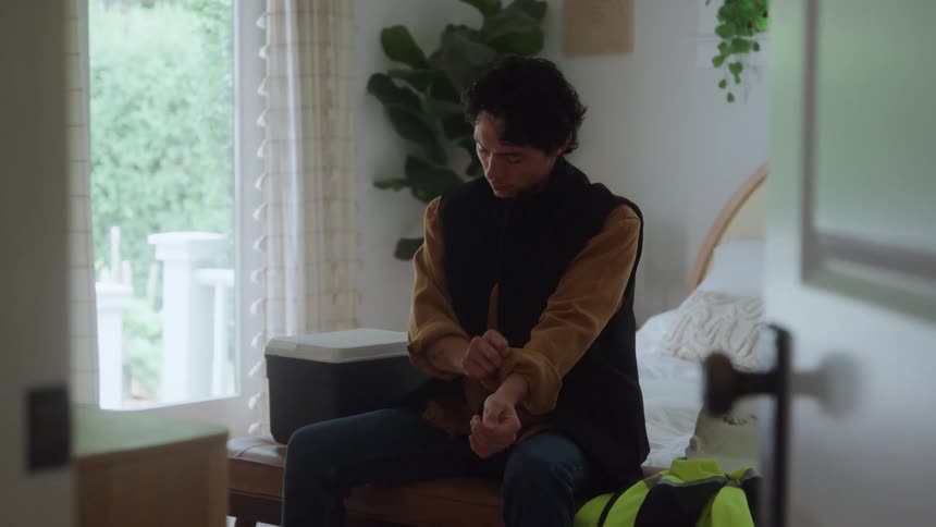 A man is sitting on a bed pulling at the sleeve of his yellow coat
