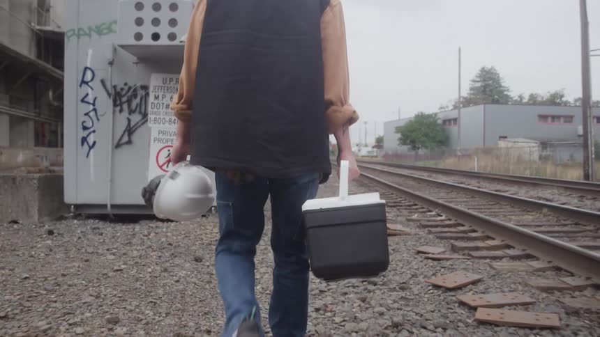 A person wearing blue jeans a light brown longsleeved shirt and a black vest is seen from behind walking away down the gravel between parallel railroad tracks carrying a white hard hat in one hand and a black and white cooler in the other
