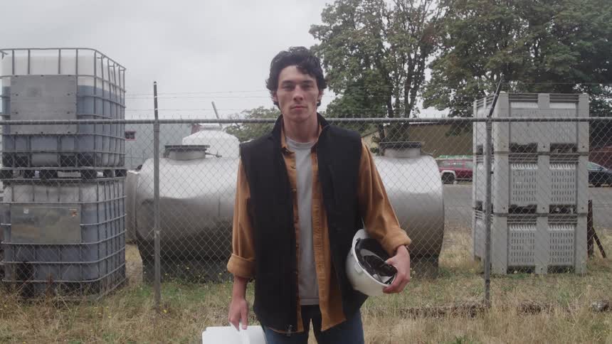 Man stands next to a fence holding his work helmet and lunch box