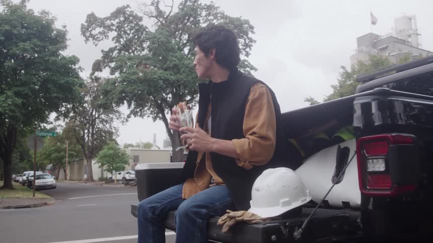 Man sitting on black pickup truck tailgate eating sandwich beside hard hats and work gloves