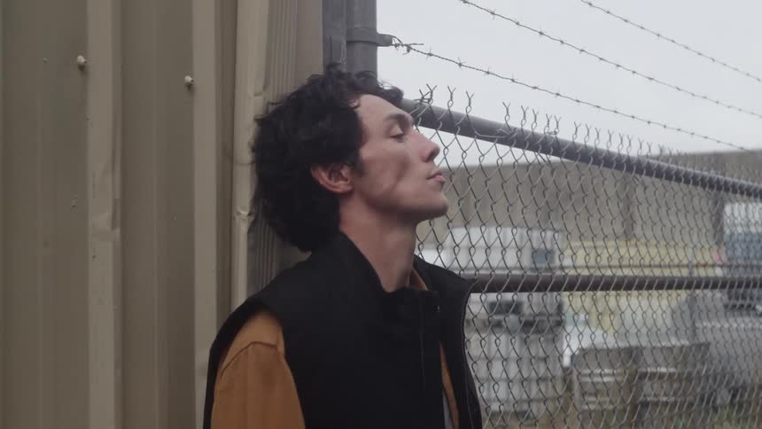 Man takes off his hard hat and leans against a wall next to a fence