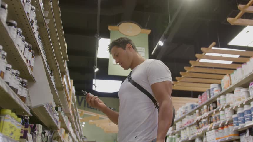 A male customer stands inside a retail shop at midday picking items from the counter with a bag strap slung across his shoulder