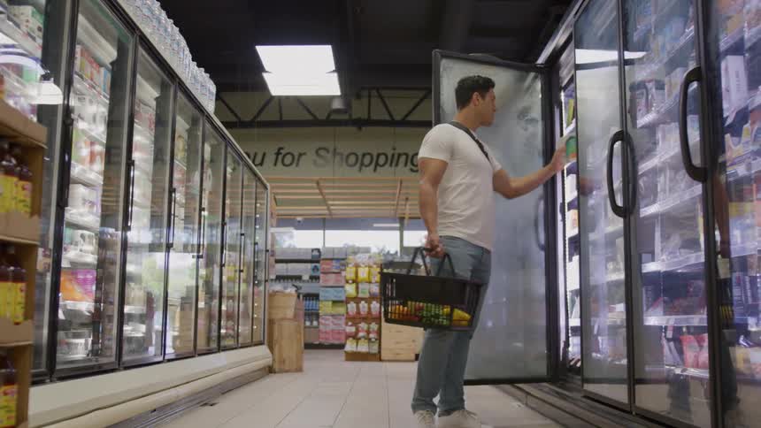 Man shopping in grocery store holding basket and inspecting item in refrigerated section