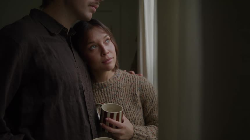 The face of a woman with her head resting on a mans chest holding a mug and looking out the window