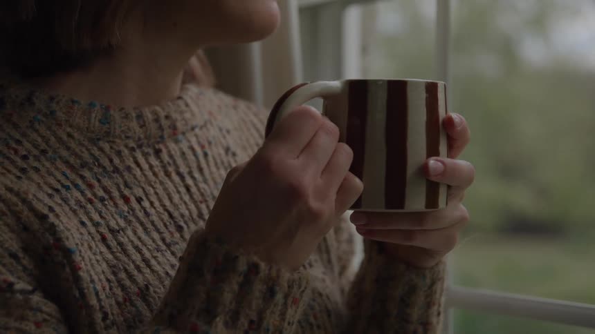 Person in sweater holds striped mug near window