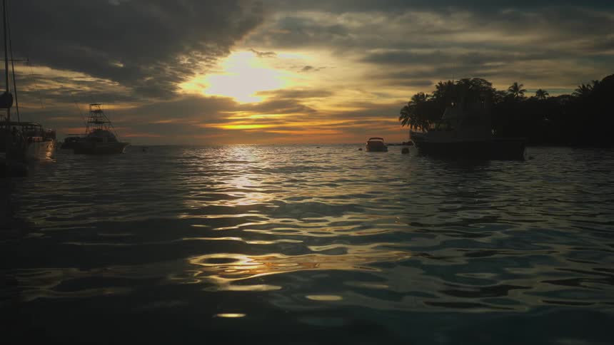 An intense orange and yellow sunset bursts through dark dramatic clouds casting a strong golden reflection across the gently rippling ocean water with fishing and pleasure boats anchored in the shadowy foreground