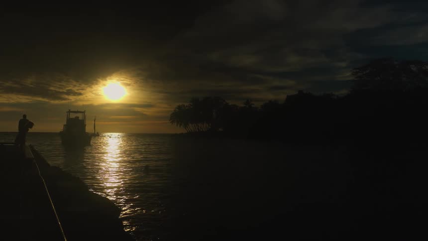 Two poeple walking on the dock as a ship sails on the water and the sun glows through clouds on the horizon