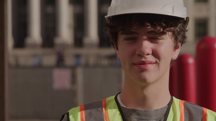 A young man stands at a construction site at midday wearing a hard hat and a green vest