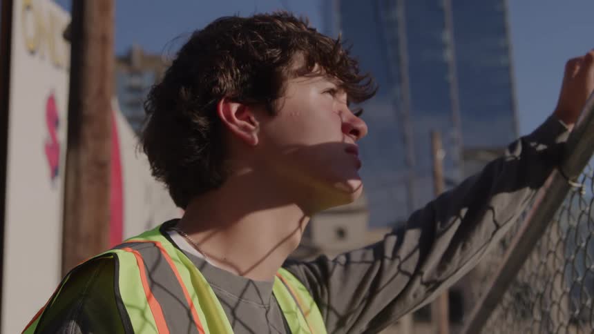 Person in reflective vest leaning on fence looking upward in urban setting with buildings in background