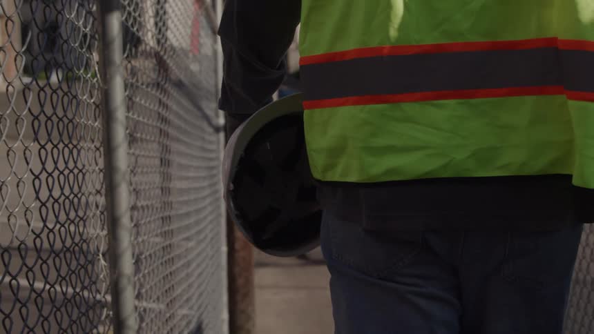Person in reflective vest walks through worksite holding a hard hat