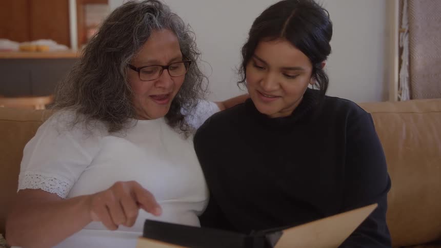 Two women an older one with long gray hair and glasses pointing to a photo and a younger one with dark hair leaning in are sitting together on a brown couch and looking happily at an open photo album