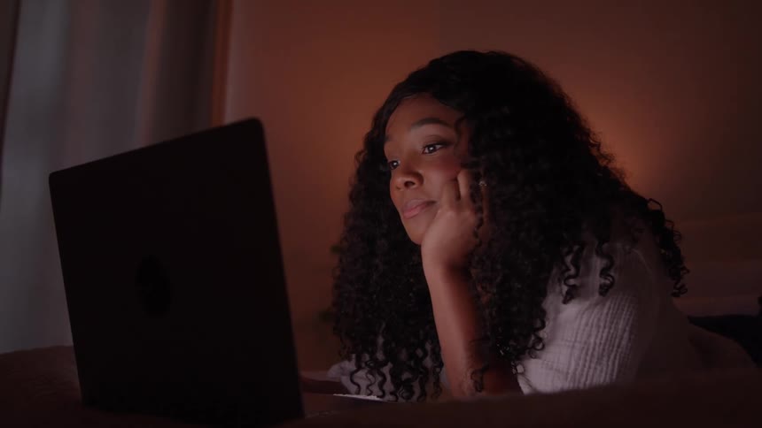 Person lying on bed looks at laptop screen in dimly lit room