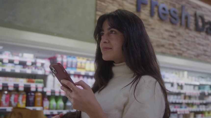 A young darkhaired woman wearing a creamcolored turtleneck holds a phone and looks thoughtfully while standing in the refrigerated section of a brightly lit grocery store