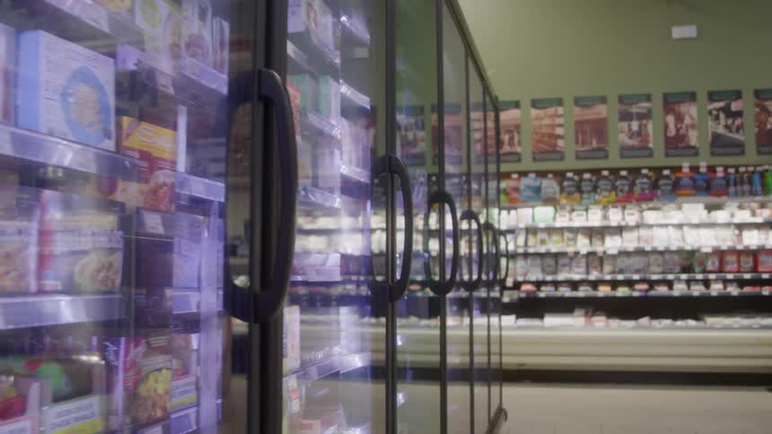 A long row of glassfronted freezer doors in a grocery store displays an array of frozen products receding into the distance against a pale green wall decorated with various promotional posters