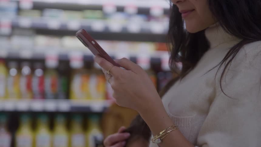 Woman stands in a store scrolling through her phone with one hand on her child