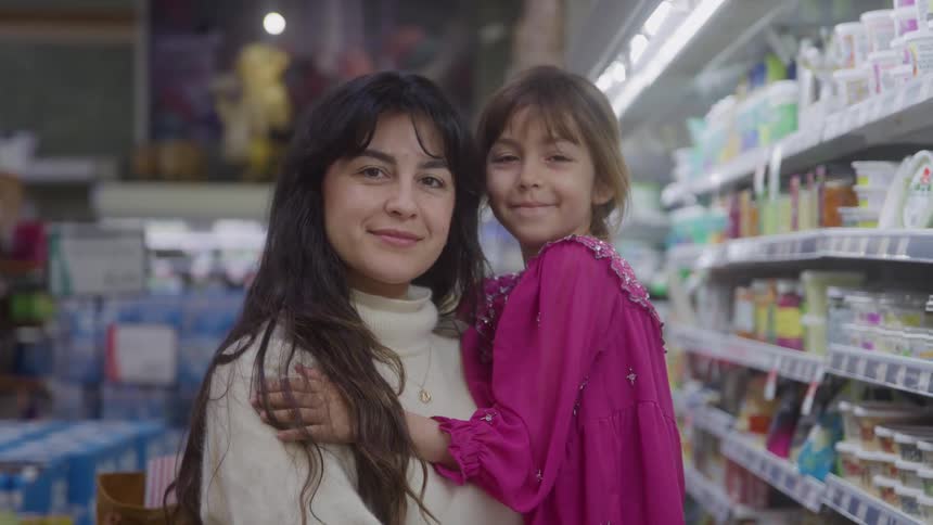 A woman wearing a lightcolored sweater is smiling and embracing a young girl in a bright pink dress as they both stand in the aisle of a brightly lit grocery store and look directly at the camera