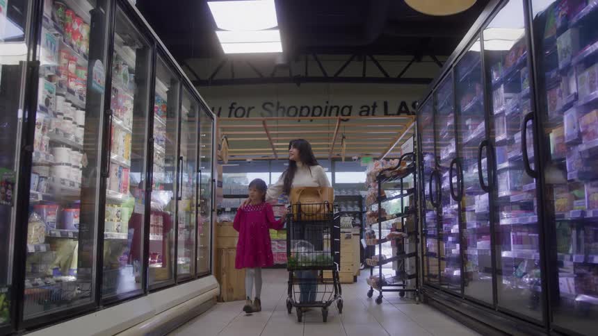 Woman pushes a cart and walks through a grocery store with her daughter looking at products in the coolers