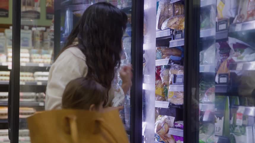 A young mother selects items from the cold section of a retail store at midday while her toddler son stands beside her