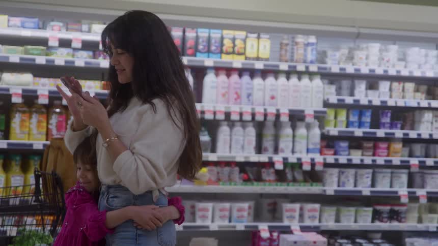Mother checks phone as daughter hugs her in grocery aisle