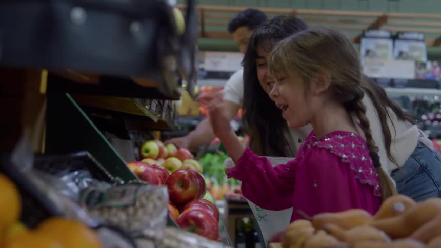 A woman and a young girl are standing at a refrigerated display in a grocery store where the woman points at and smiles at the red and green apples as the girl handles an apple and holds a plastic bag and a man on the back picking fruits too
