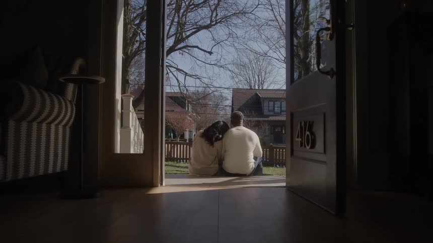 A man and a woman are sitting on the porch of a house with the door open looking straight ahead and with their backs turned