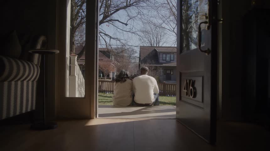 Indoor view shows two people seated on steps gazing at peaceful neighborhood