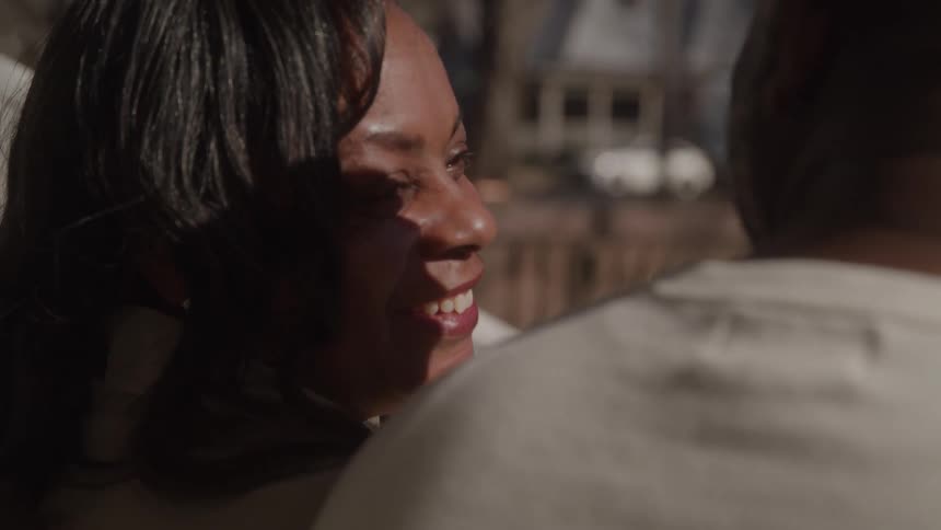 A smiling middleaged woman with dark hair beams genuinely while looking towards an unseen person whose side of his face is visible in the foreground