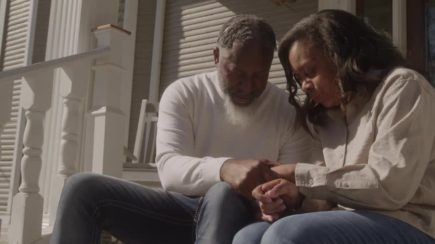 Two people seated on porch steps holding hands and bowing heads in a moment of prayer