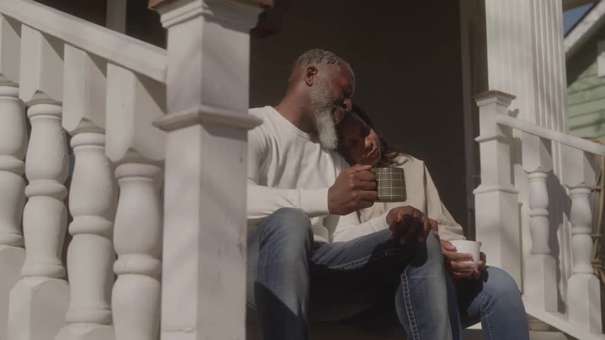 An older man with a white beard and an older woman are sitting closely together on the steps of a white wooden porch both holding mugs and sharing an intimate moment in the bright sun