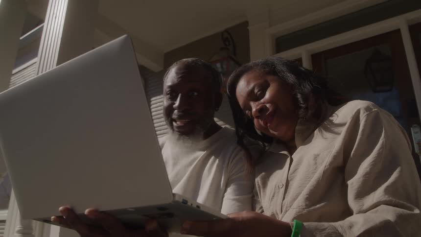 An older man and a middleaged woman are joyfully looking and waving at and interacting with a silver laptop screen held between them sitting on a porch beneath the bright sunlight
