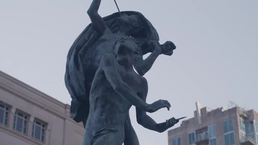 A dramatic bronze statue of two figures stands in a city with modern buildings visible in the background