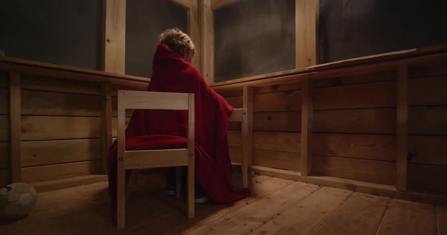 Little boy wrapped in red blanket is sitting at desk and drawing