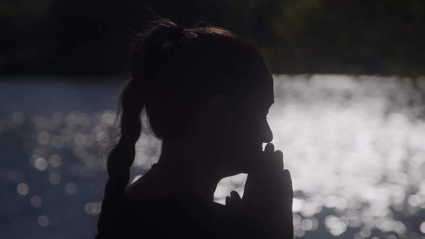 The silhouette of a womans head with her hands over her mouth and behind her a body of water with sunlight illuminating its surface