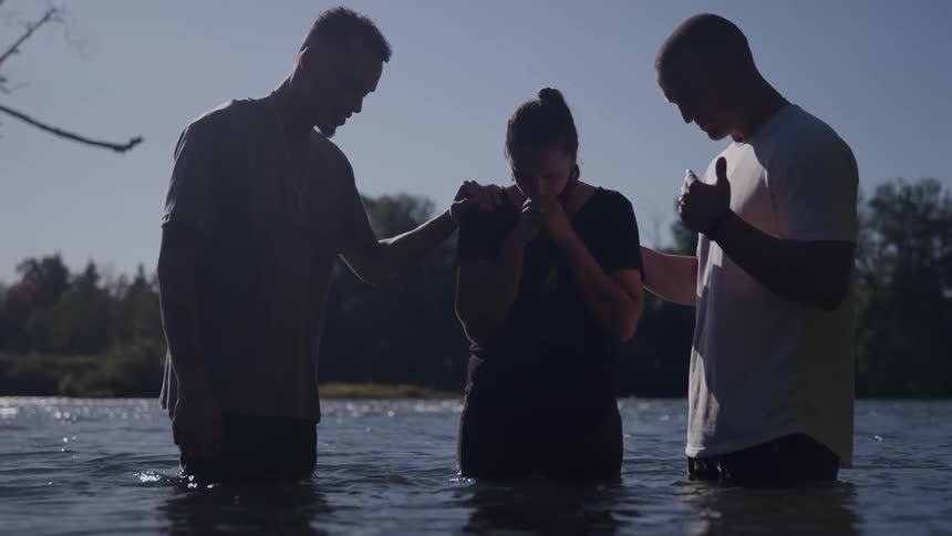 Three people standing in shallow river take part in ritual with supportive gestures