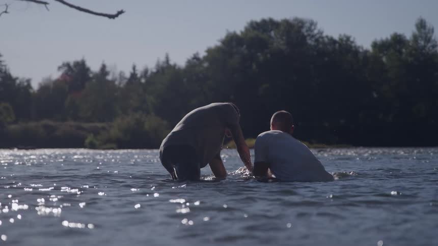 Two men holding a woman and submerging her in a shallow body of water then pulling her to the surface