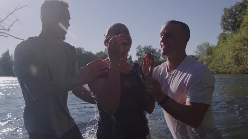 Two men one in a white shirt and one in a dark shirt stand in a sunny lake and joyfully immerse and then raise a smiling woman from the water